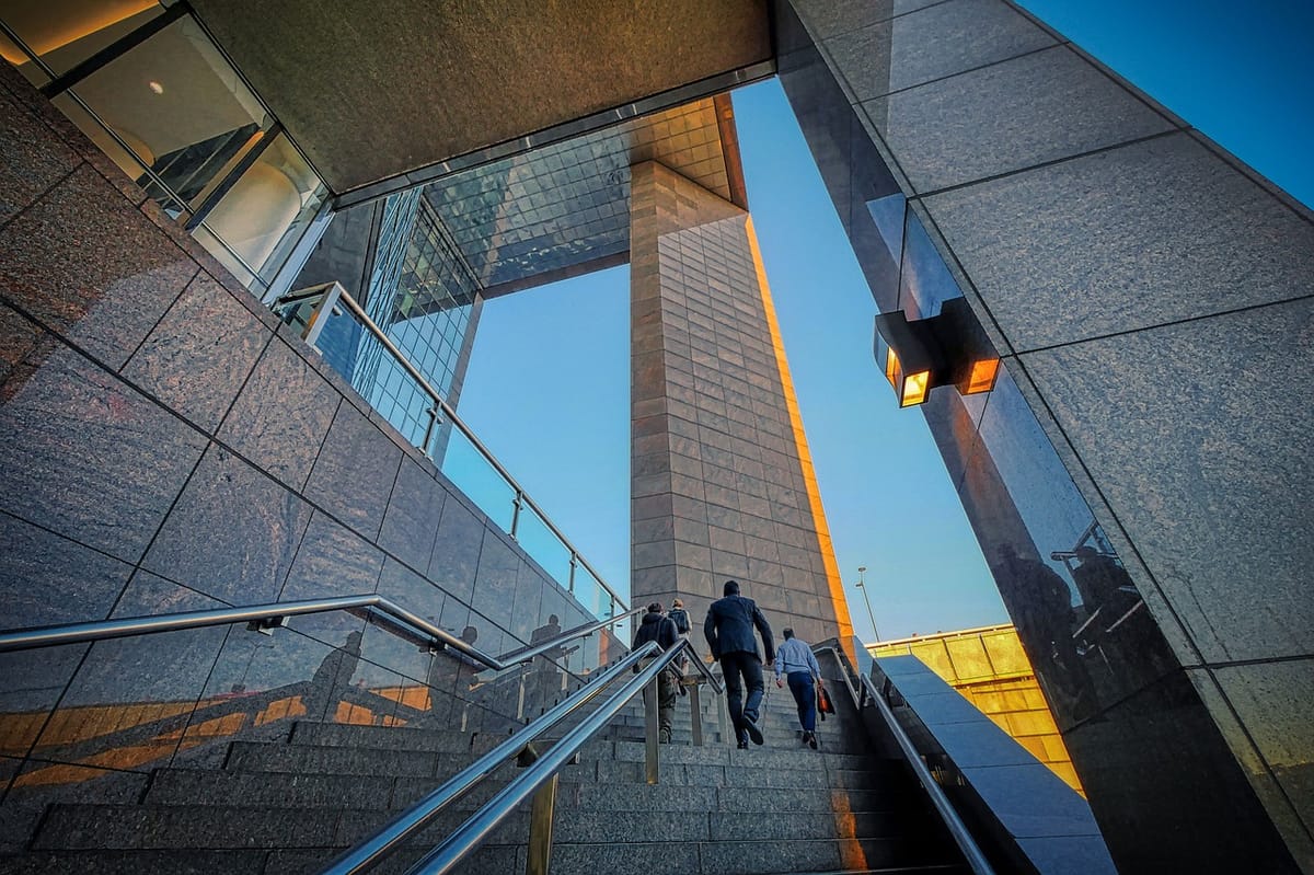 Two businessmen climbing stairs inside a glass office tower, representing incremental steps toward a successful business exit